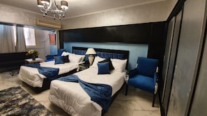 Una habitación de hotel con dos camas, un sillón azul, una araña y una ventana con cortinas.