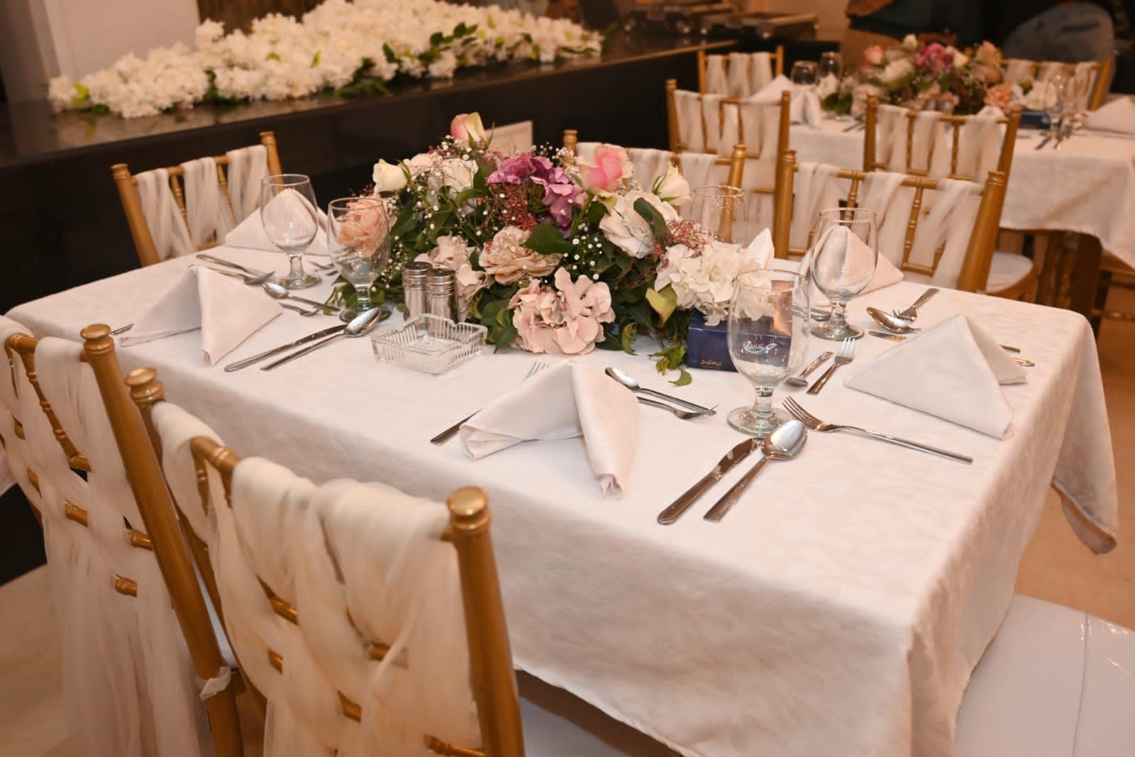 Uma mesa de banquete posta com porcelana requintada, talheres e taças de cristal, rodeada por cadeiras de madeira com toalhas brancas.