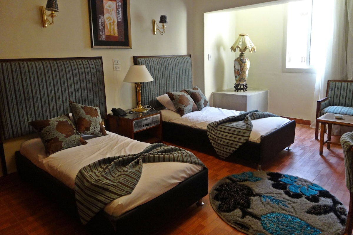 Una habitación de hotel con dos camas, una zona de estar, un ventanal y una lámpara.