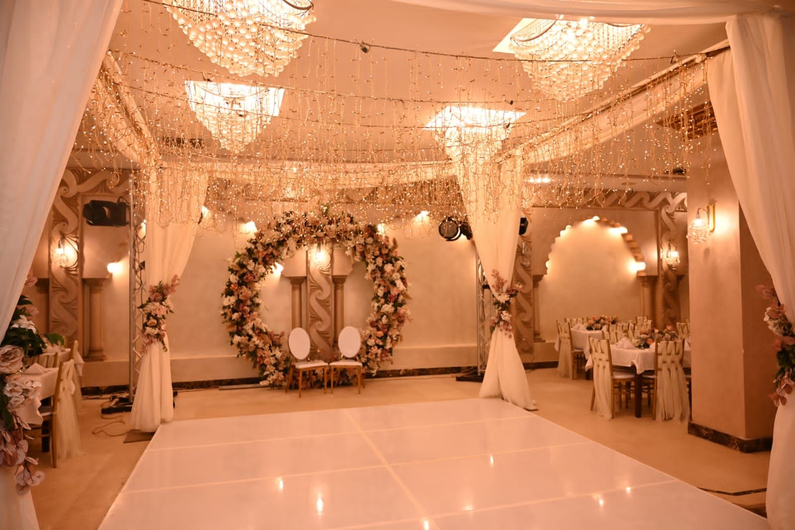 A wedding reception area with a white dance floor, floral decorations, and chandeliers.