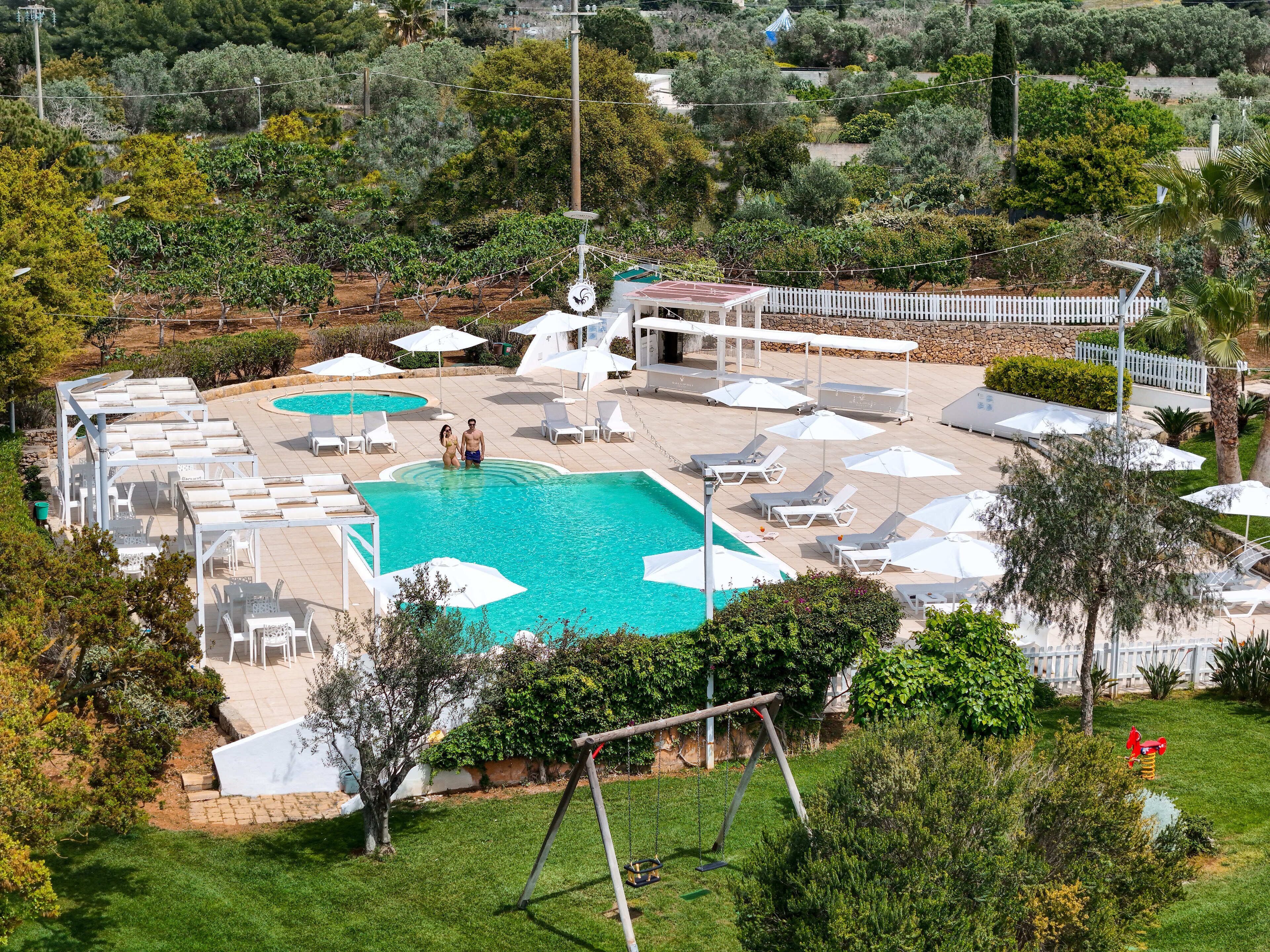 Una gran piscina al aire libre con múltiples zonas de descanso y sombrillas, rodeada de follaje y una valla blanca.