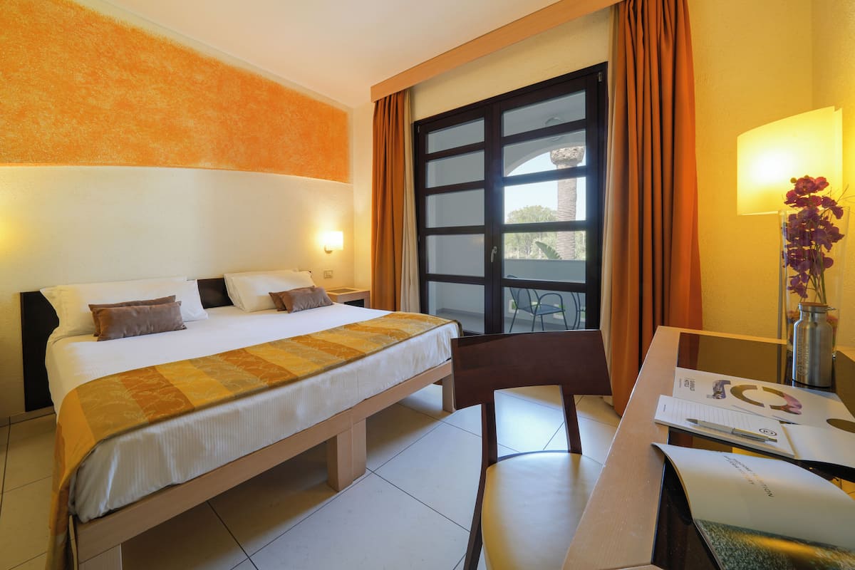 A hotel room with a bed, a desk with a chair, a window with curtains, and a vase with flowers.