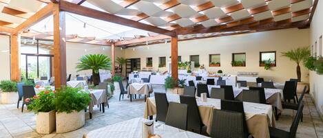 An outdoor dining area with tables and chairs, potted plants, and a ceiling with wooden beams.