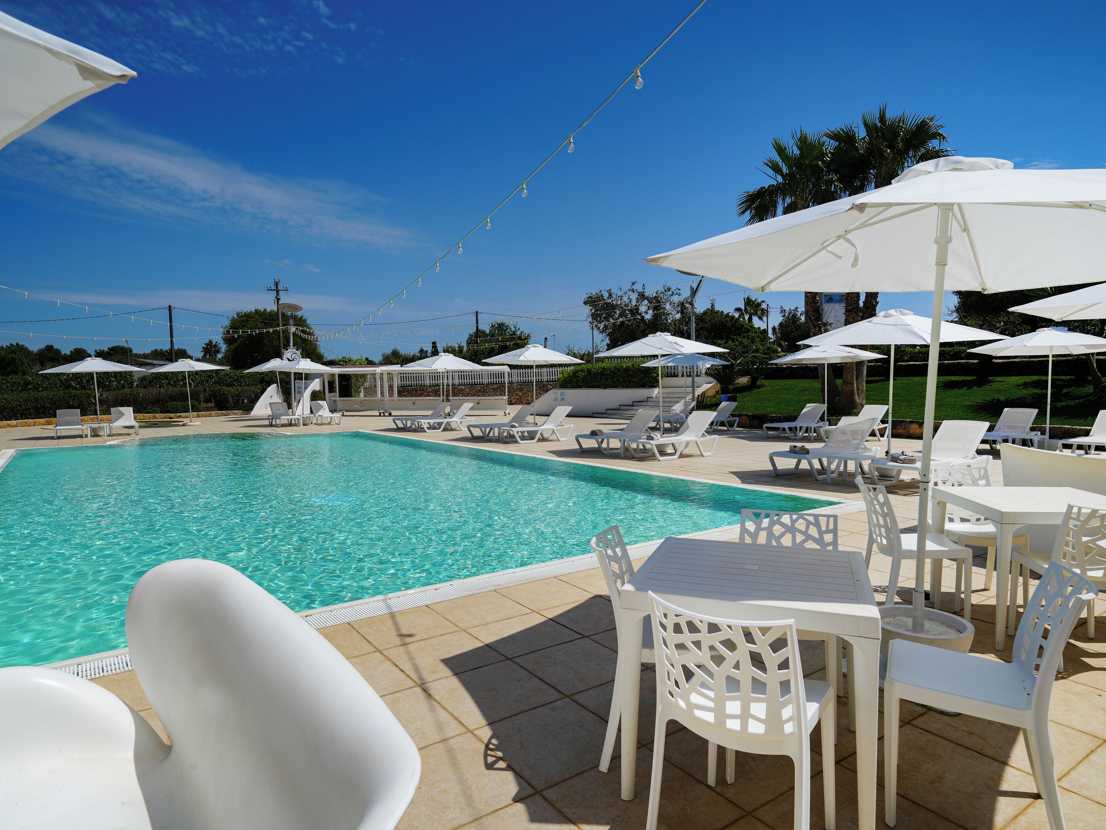 A pool area with white lounge chairs and umbrellas, a tiled deck, and a clear blue sky.