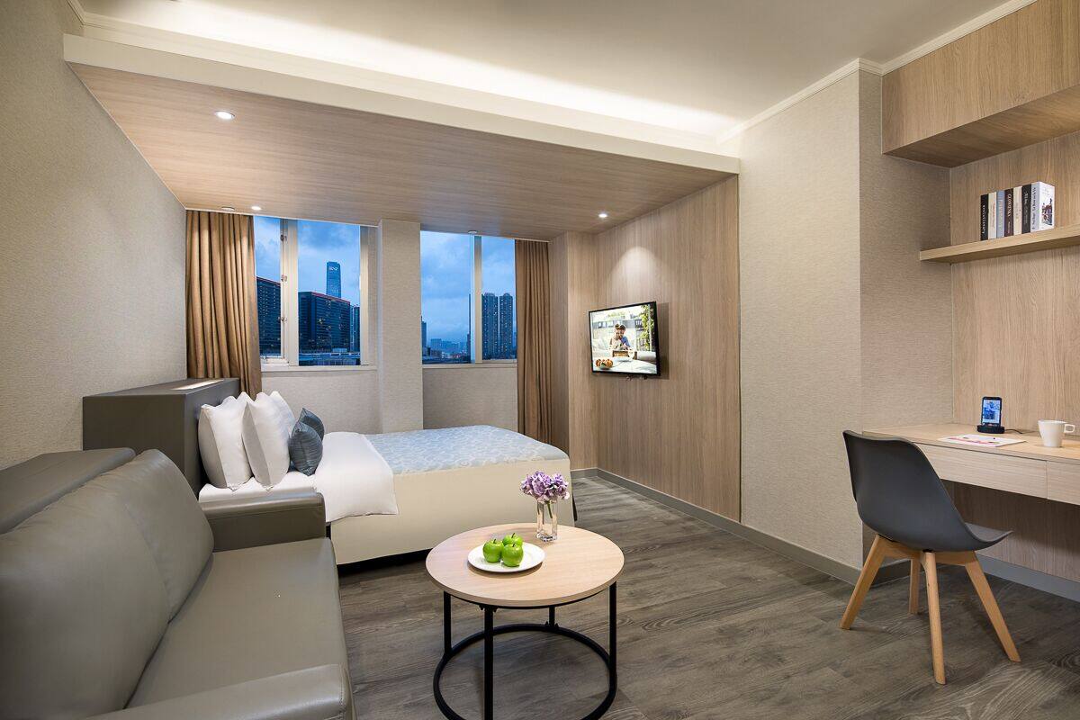 A modern hotel room with a bed, a desk, a chair, a small table with a vase and apples, and a city view through large windows.