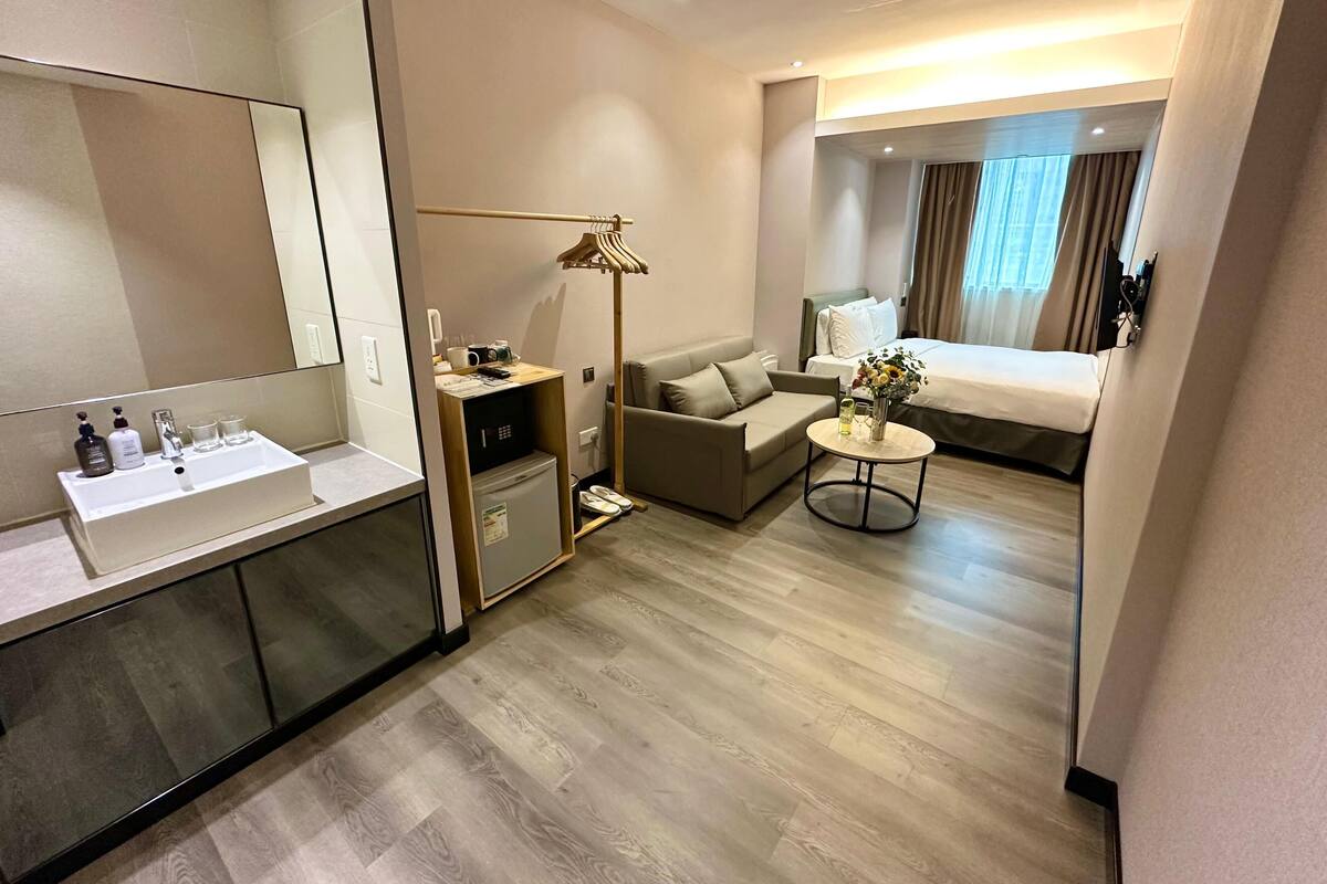 A hotel room with a large mirror, a sink, a small desk, a sofa, a round table with a vase, and a bed with curtains.