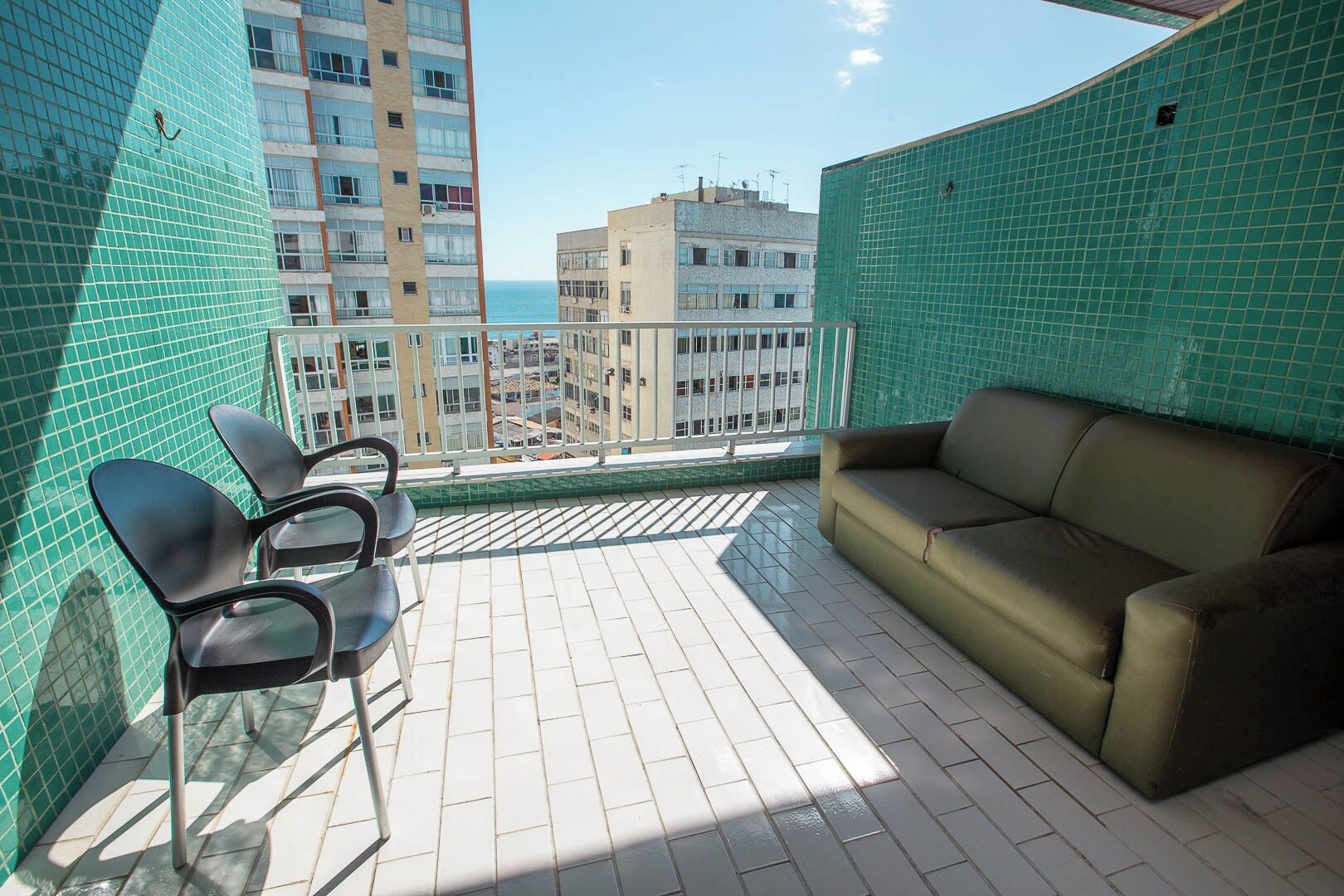 Ein Balkon mit grünen Fliesen, einem beigen Sofa und zwei schwarzen Stühlen.