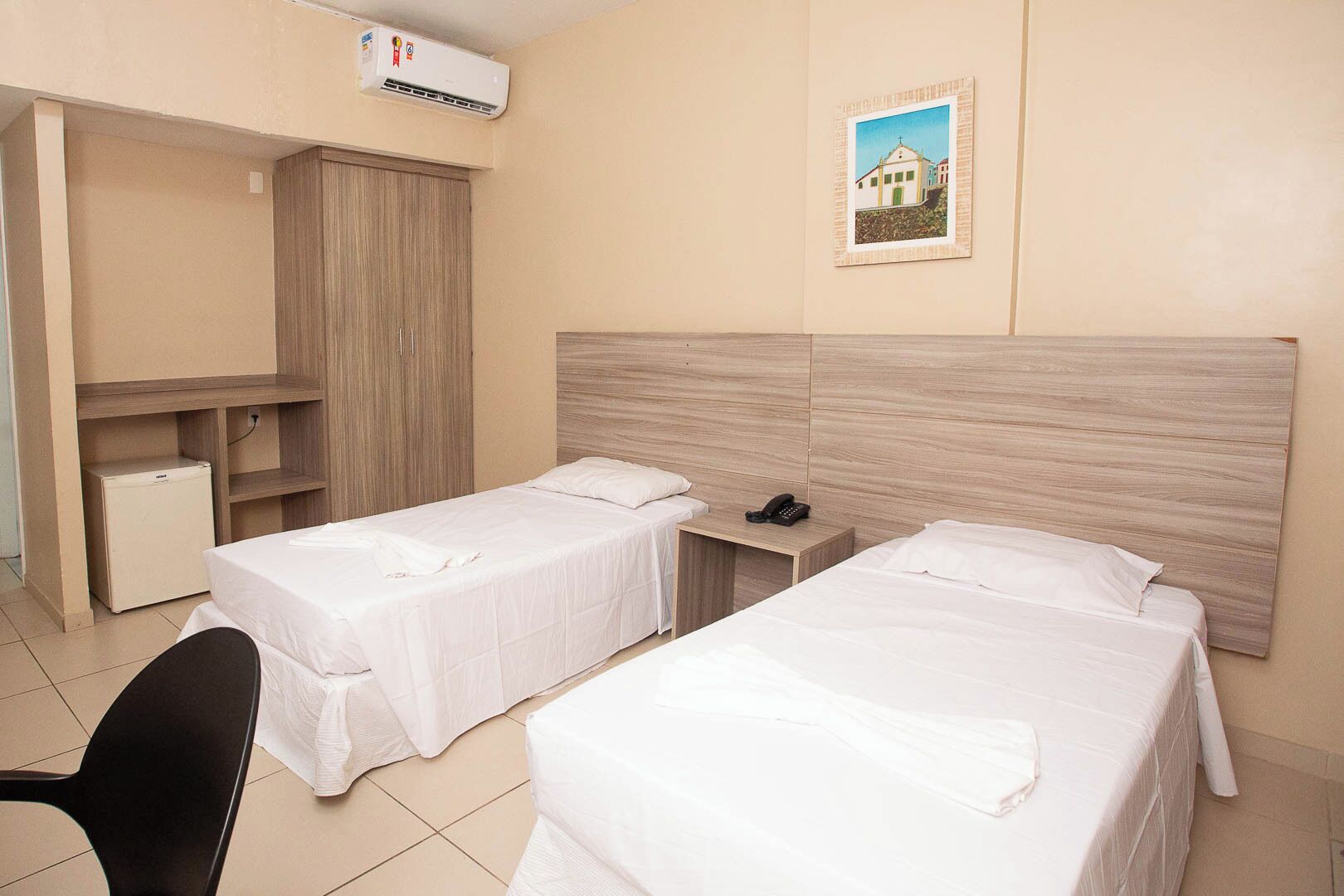 Quarto de hotel com duas camas de solteiro, uma escrivaninha, uma cadeira, um guarda-roupa e um pequeno frigorífico.