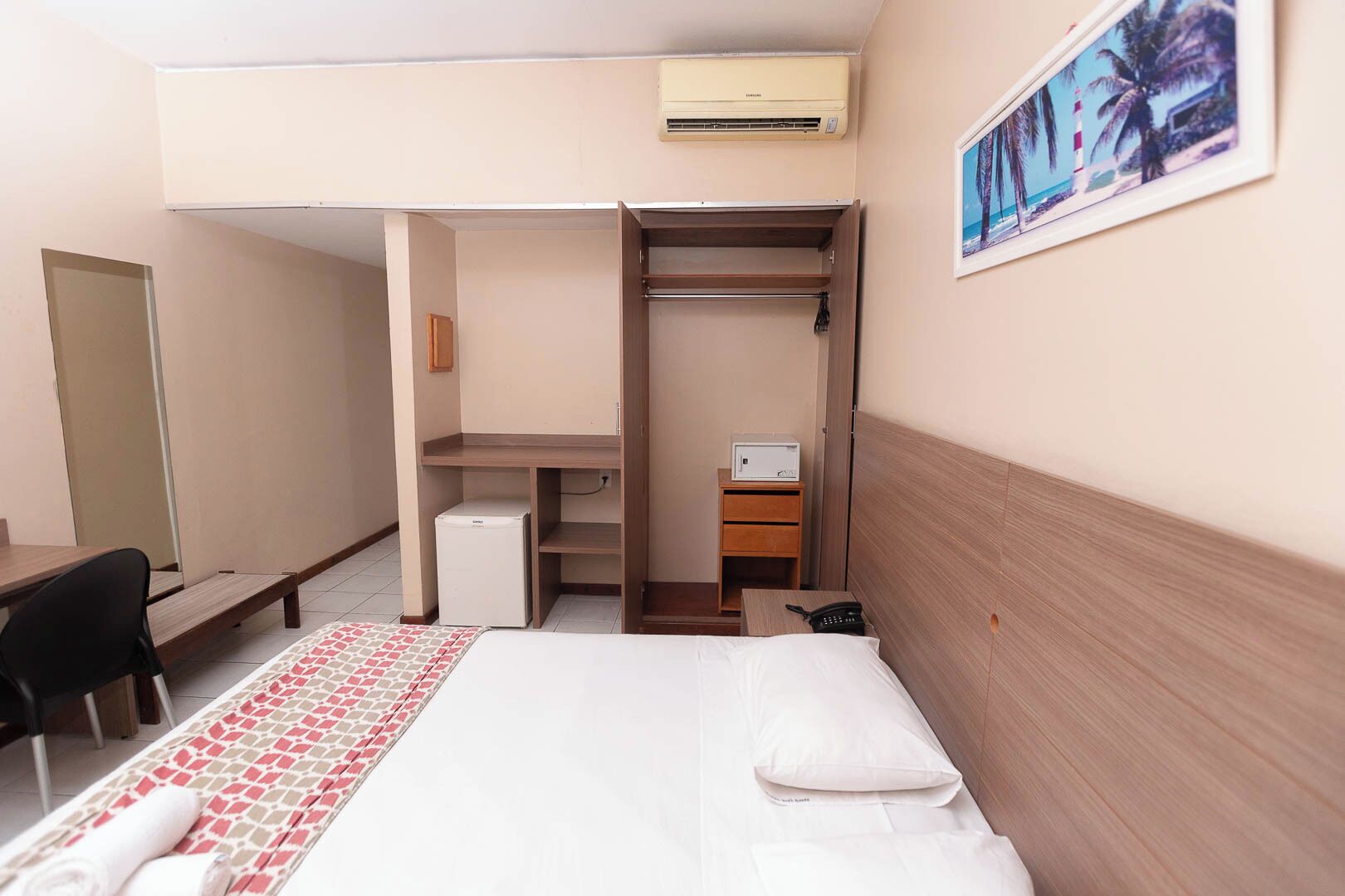 Ein Hotelzimmer mit Bett, Schreibtisch, Stuhl, Kleiderschrank, kleinem Kühlschrank und einem Bild an der Wand.