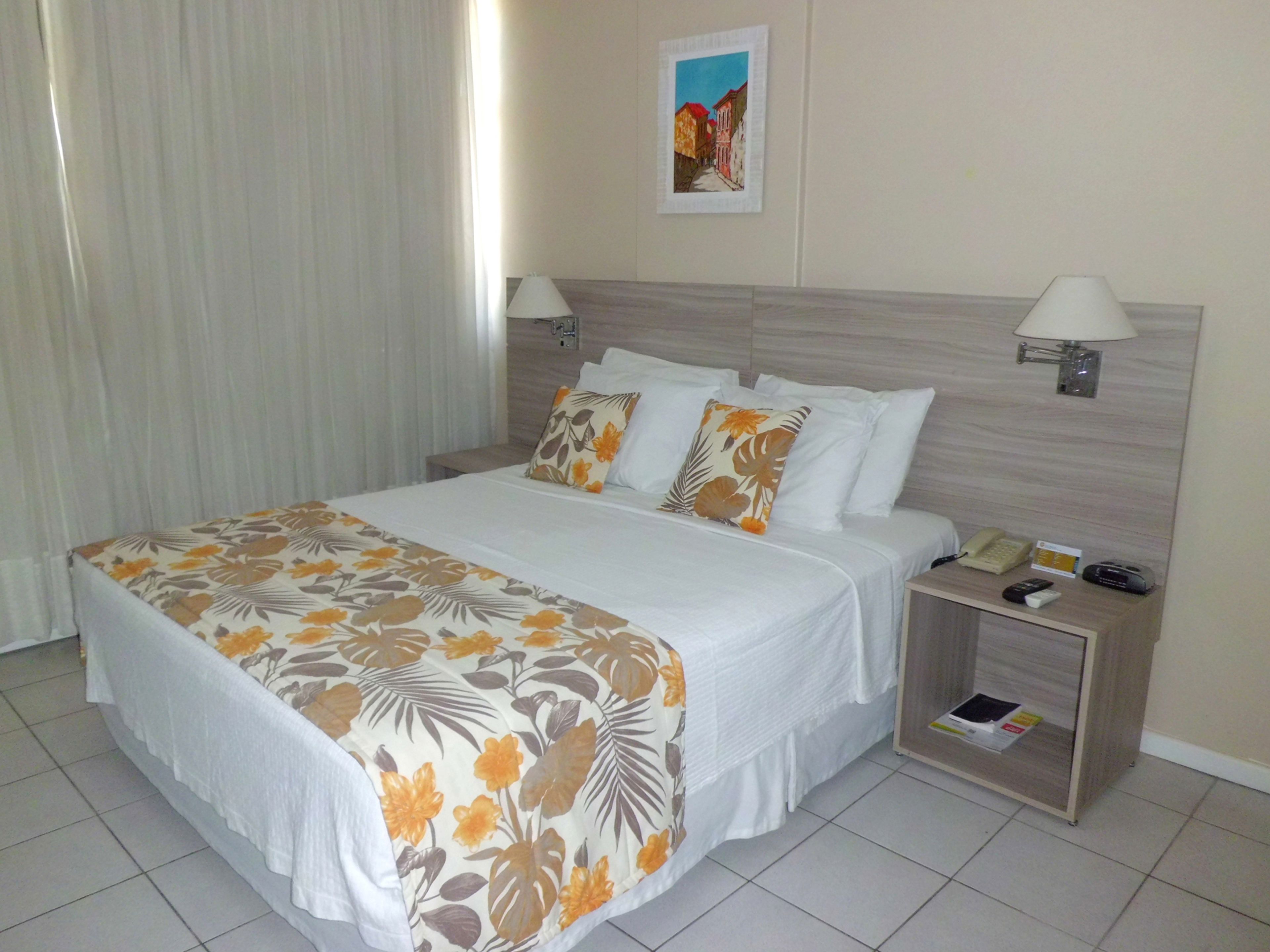 A hotel room with a large bed, two bedside tables, a nightstand, a lamp, a painting on the wall, and a window with curtains.