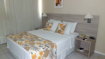 Quarto de hotel com uma cama grande, duas mesas de cabeceira, um criado-mudo, uma luminária, um quadro na parede e uma janela com cortinas.