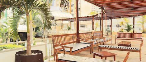 An outdoor patio with wooden furniture, including benches and a table, surrounded by potted plants and a pergola structure.