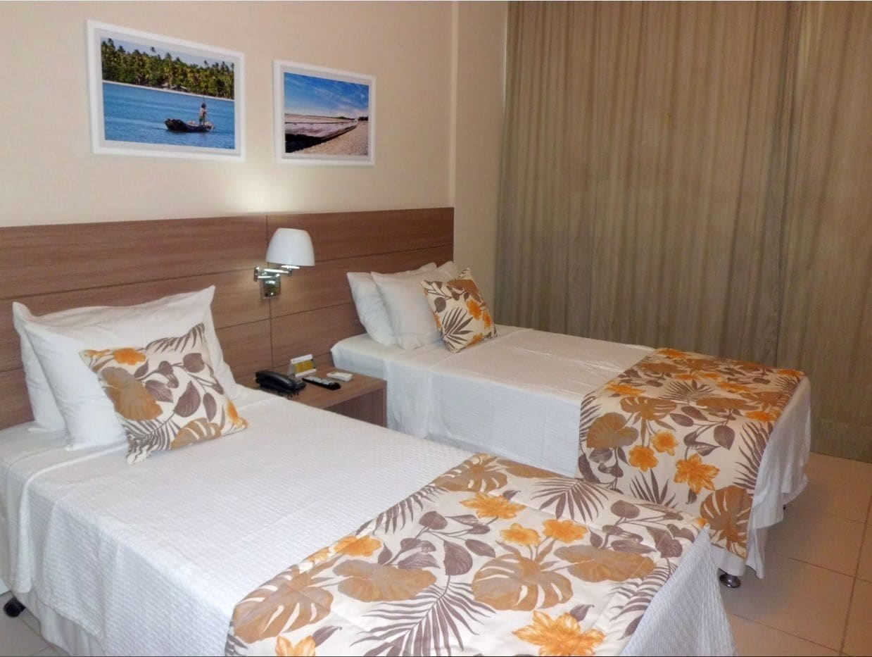 A hotel room with two single beds, a nightstand with a lamp, and two framed pictures on the wall.