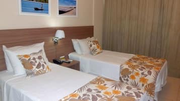 Quarto de hotel com duas camas de solteiro, uma mesa de cabeceira com abajur e duas fotos emolduradas na parede.