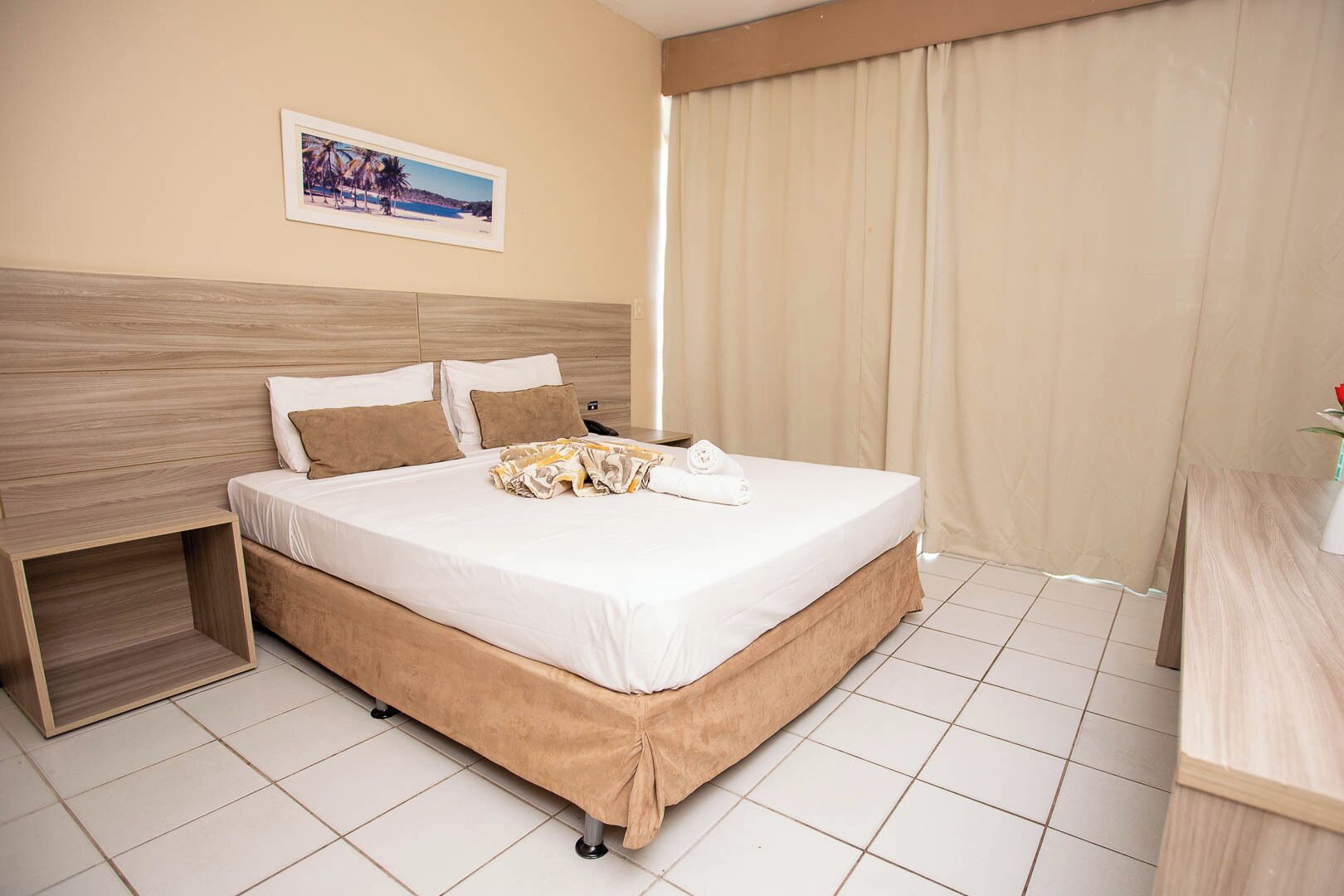 A hotel room with a bed, a nightstand, a picture on the wall, and a window with curtains.