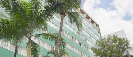 A multi-story hotel with a green and white facade, palm trees, and a paved courtyard.