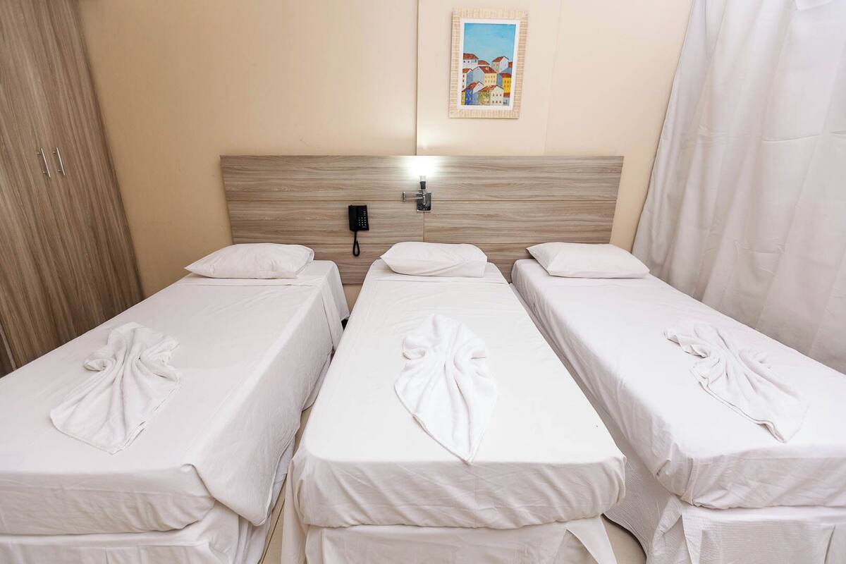 A hotel room with three single beds, each with a neatly folded towel. There is a wooden headboard, a wall-mounted phone, and a framed picture above the beds.