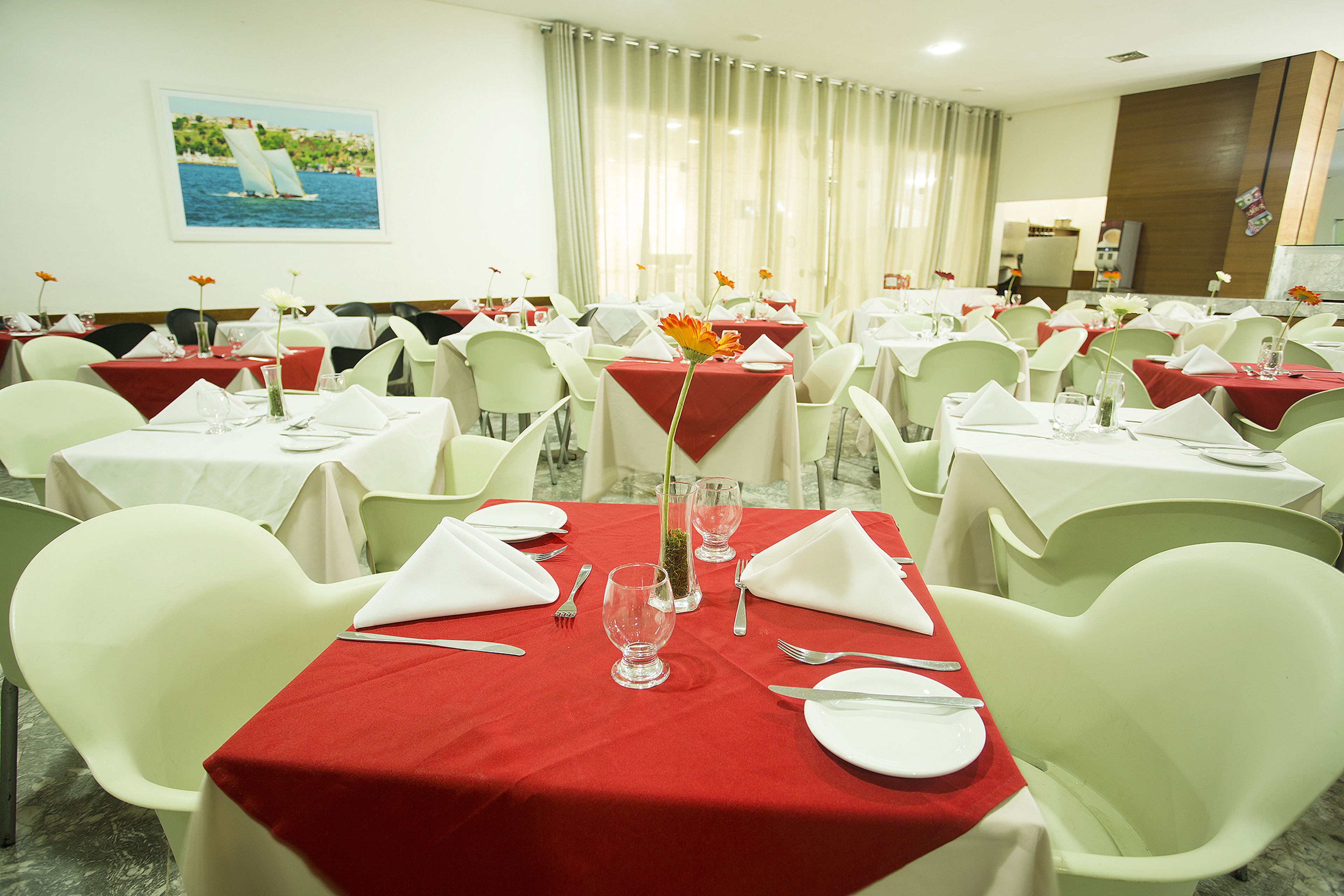 Une salle à manger avec des tables dressées, agrémentée d’une nappe rouge, de chaises blanches et offrant une vue sur la mer par la fenêtre.