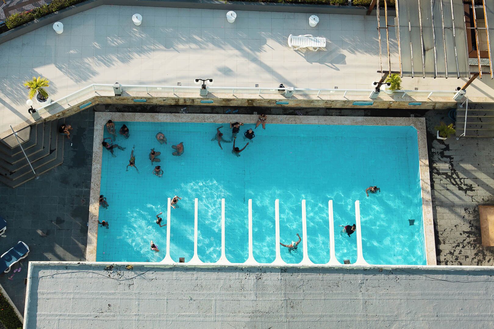 Une vue aérienne d’une piscine où des personnes nagent et prennent des bains de soleil.