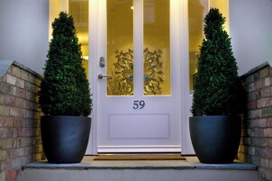 Puerta principal con el número 59, flanqueada por dos árboles en macetas y un panel de vidrio decorativo.