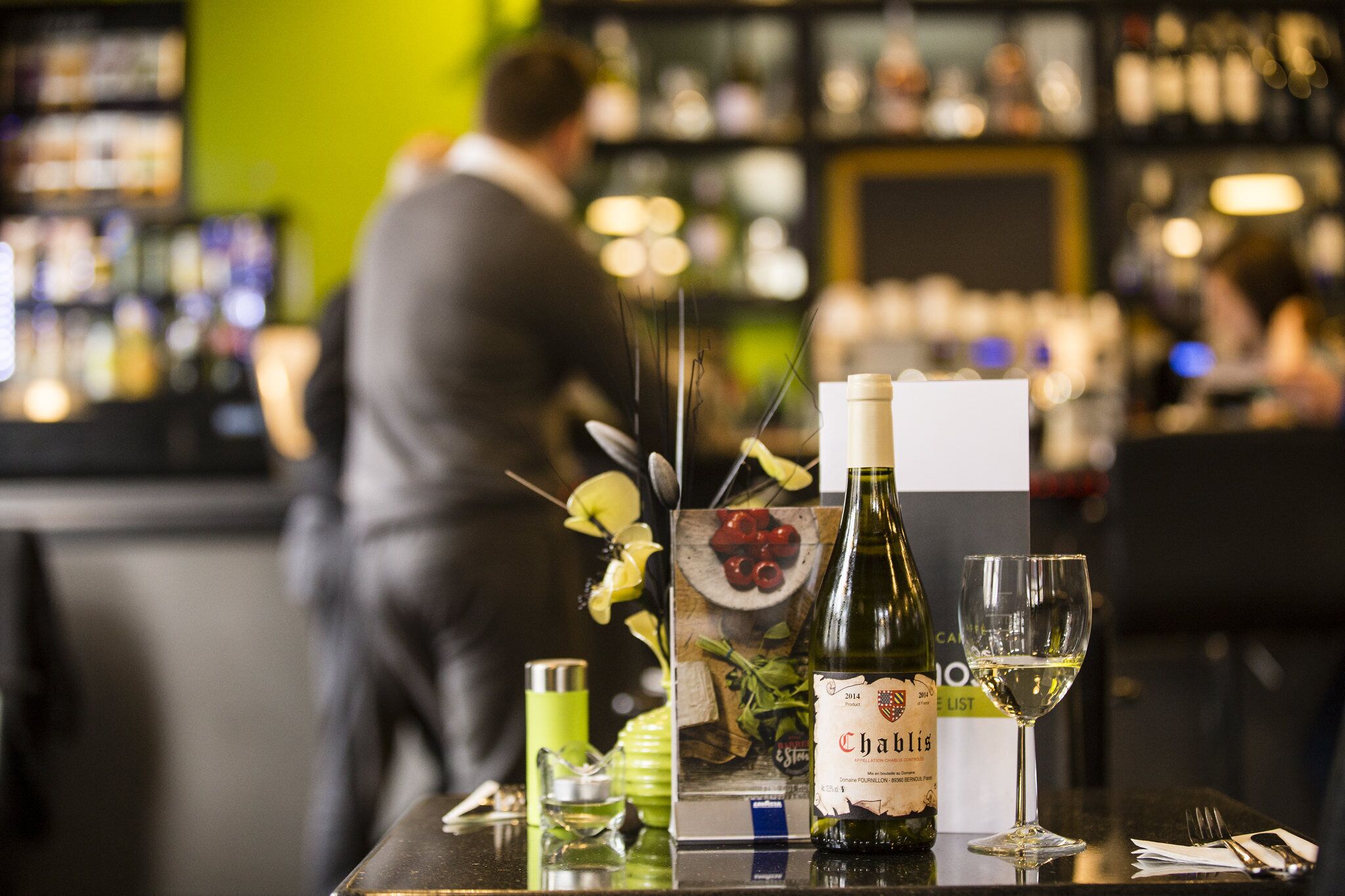 A bar setting with a bottle of Chablis wine, a glass of white wine, and a menu on the table.