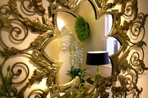 Un espejo con marco dorado ornamentado, una estatua de Buda y una planta en maceta con flores blancas.