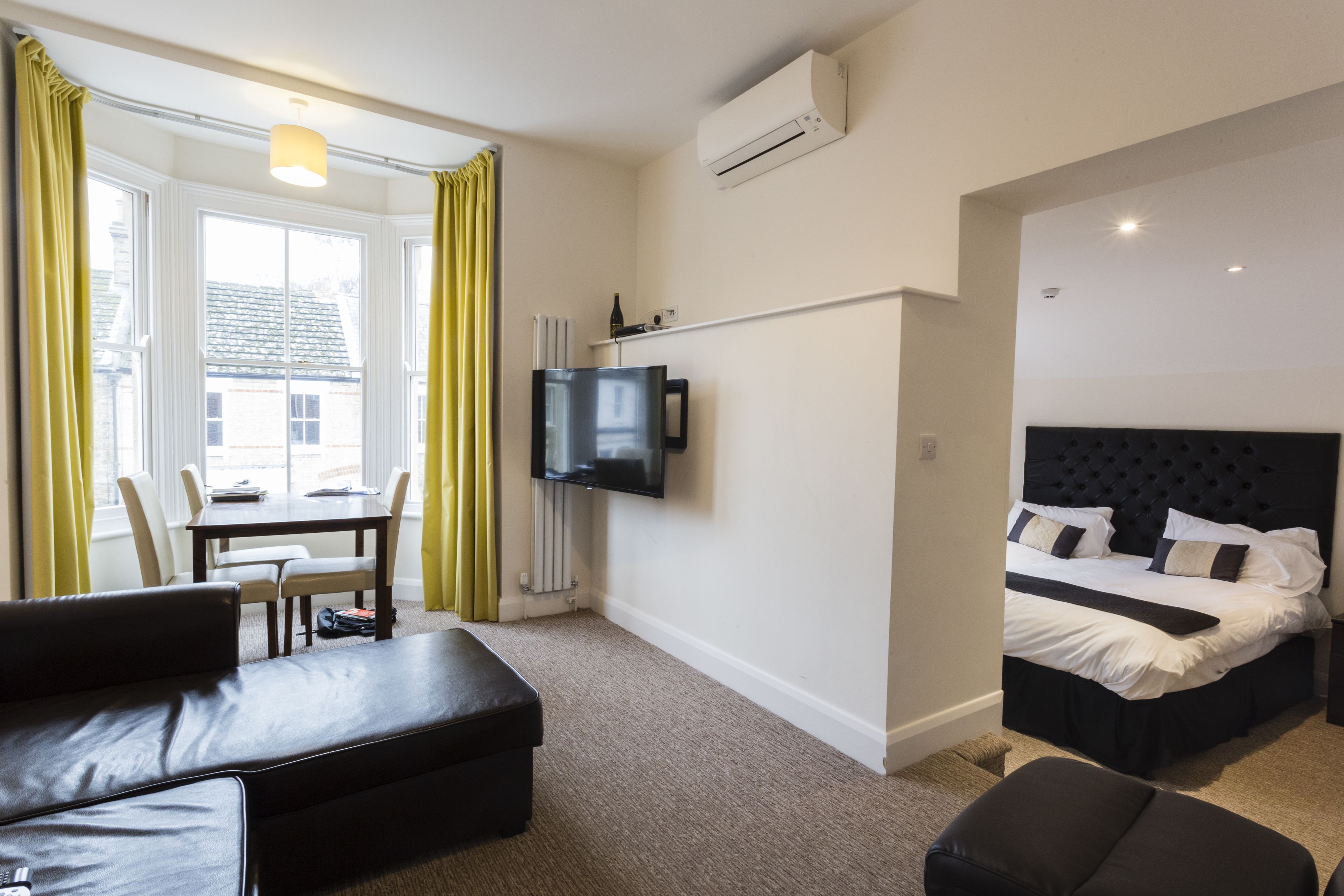 A modern hotel room with a bed, a TV, a dining area with a table and chairs, and a view of the outside.