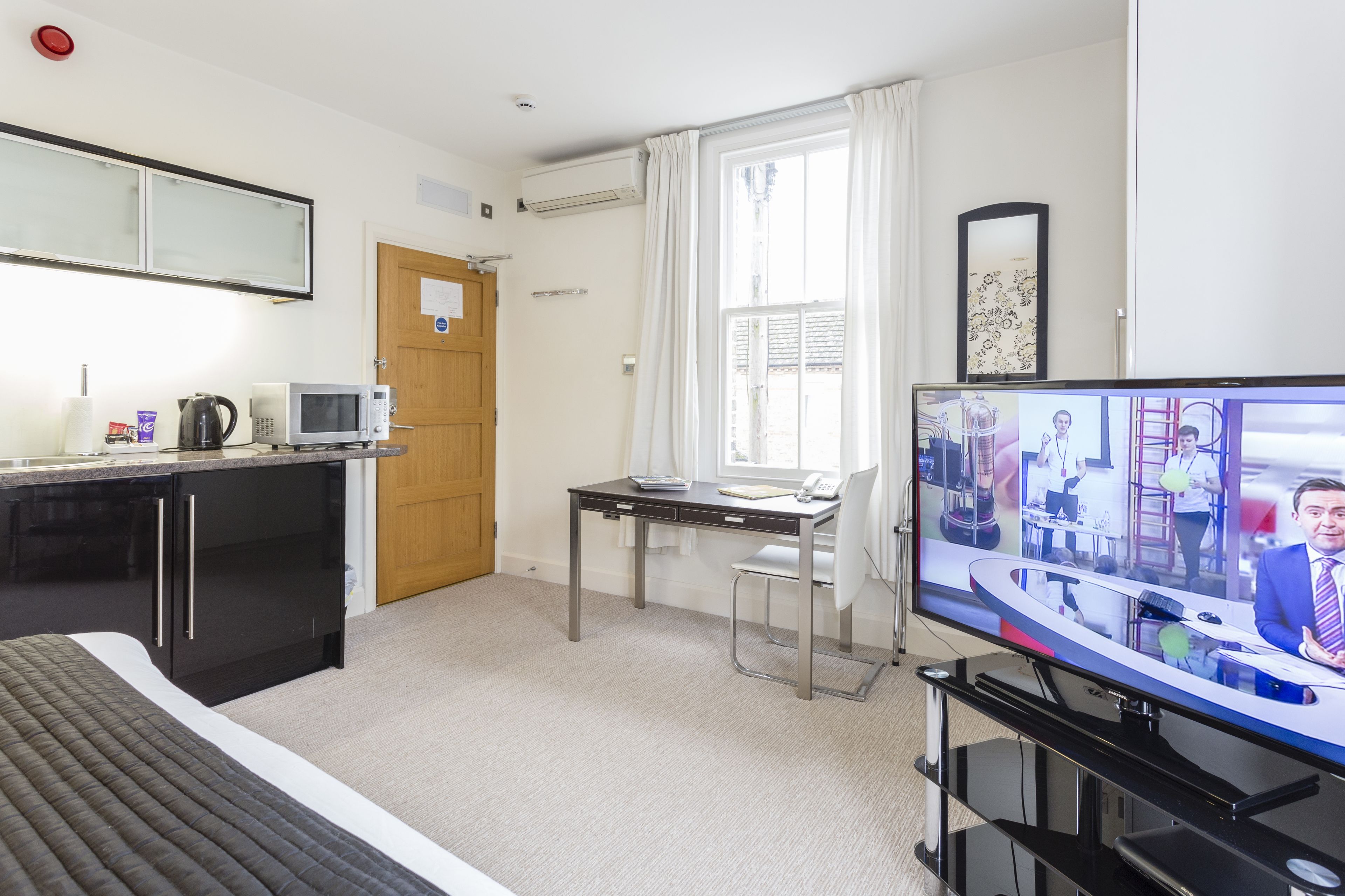 A hotel room with a bed, a desk with a computer, a TV, and a kitchenette.