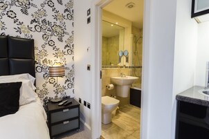 Habitación de hotel con cama, mesita de noche con lámpara, baño con lavamanos, inodoro y ducha.