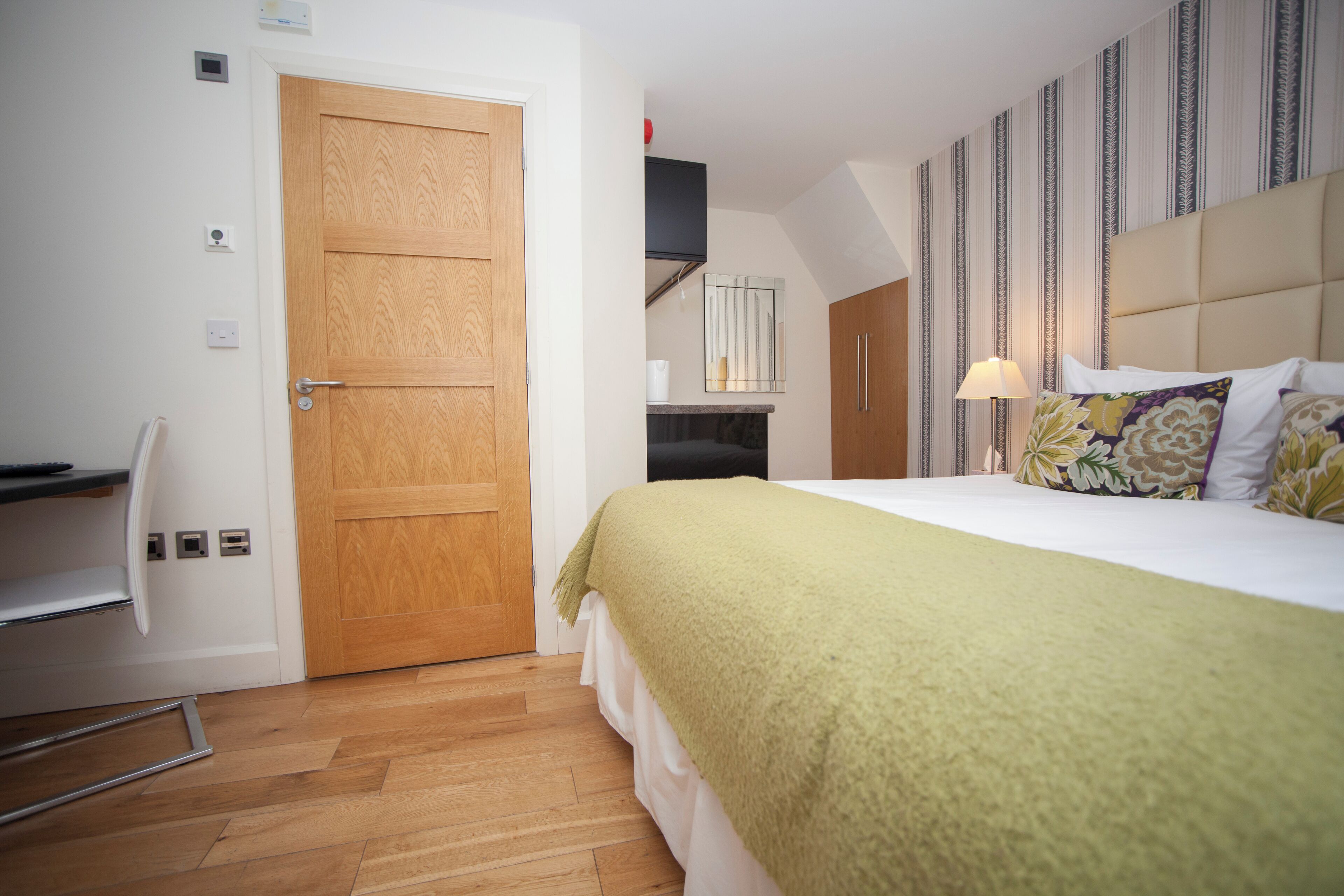 Una habitación de hotel con una puerta de madera, una cama con una manta verde, una lámpara de noche y una pared a rayas.