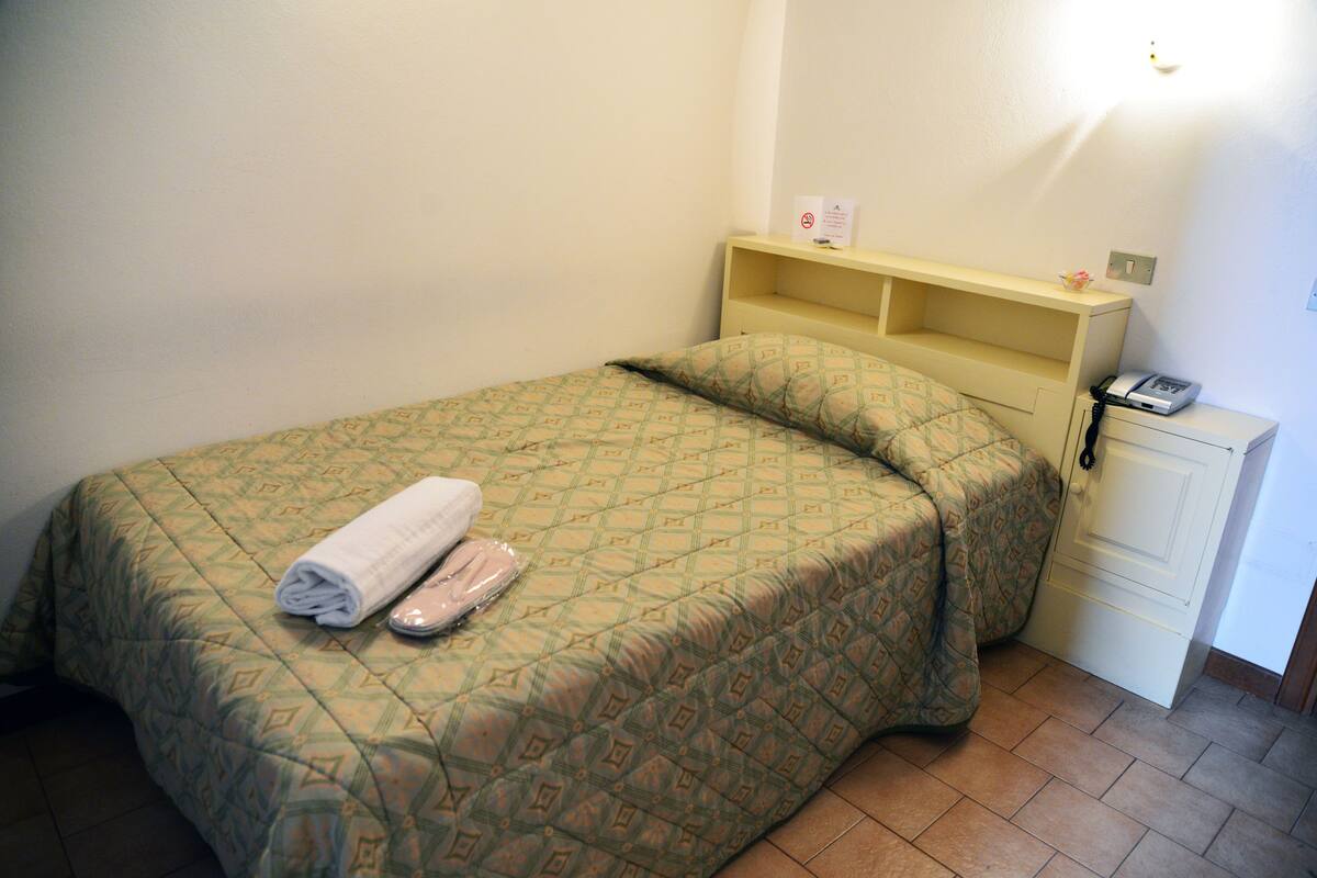 A single bed with a patterned bedspread, a rolled towel, and slippers on it. There is a nightstand with a phone and a small shelf.
