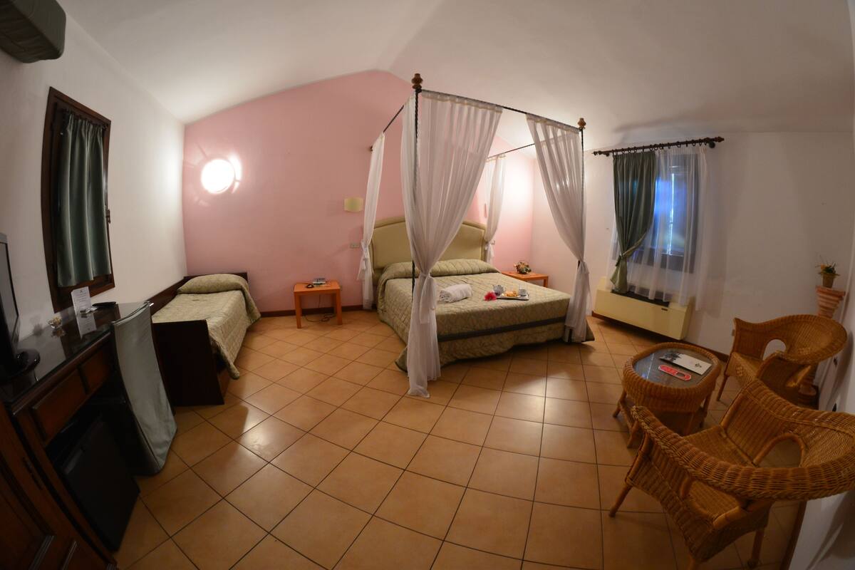 A bedroom with a four-poster bed, a small bedside table, a chair, a desk, and a window with curtains.