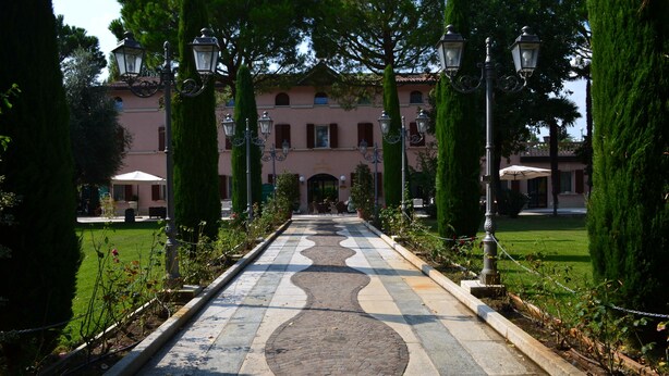A pathway leading to a building with a pink facade, flanked by tall cypress trees and ornate street lamps. - Panazza Hotel (Mordano)
