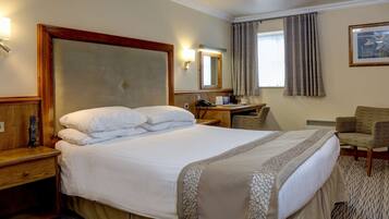 A hotel room with a large bed, a desk, a chair, a bedside table, a lamp, a telephone, and a window with curtains.