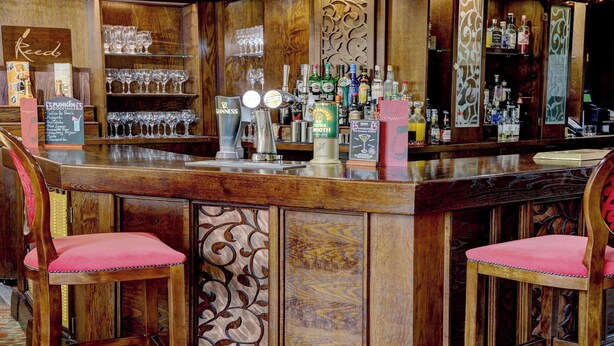 Una zona de bar con mostrador de madera, taburetes y diversas botellas y vasos.