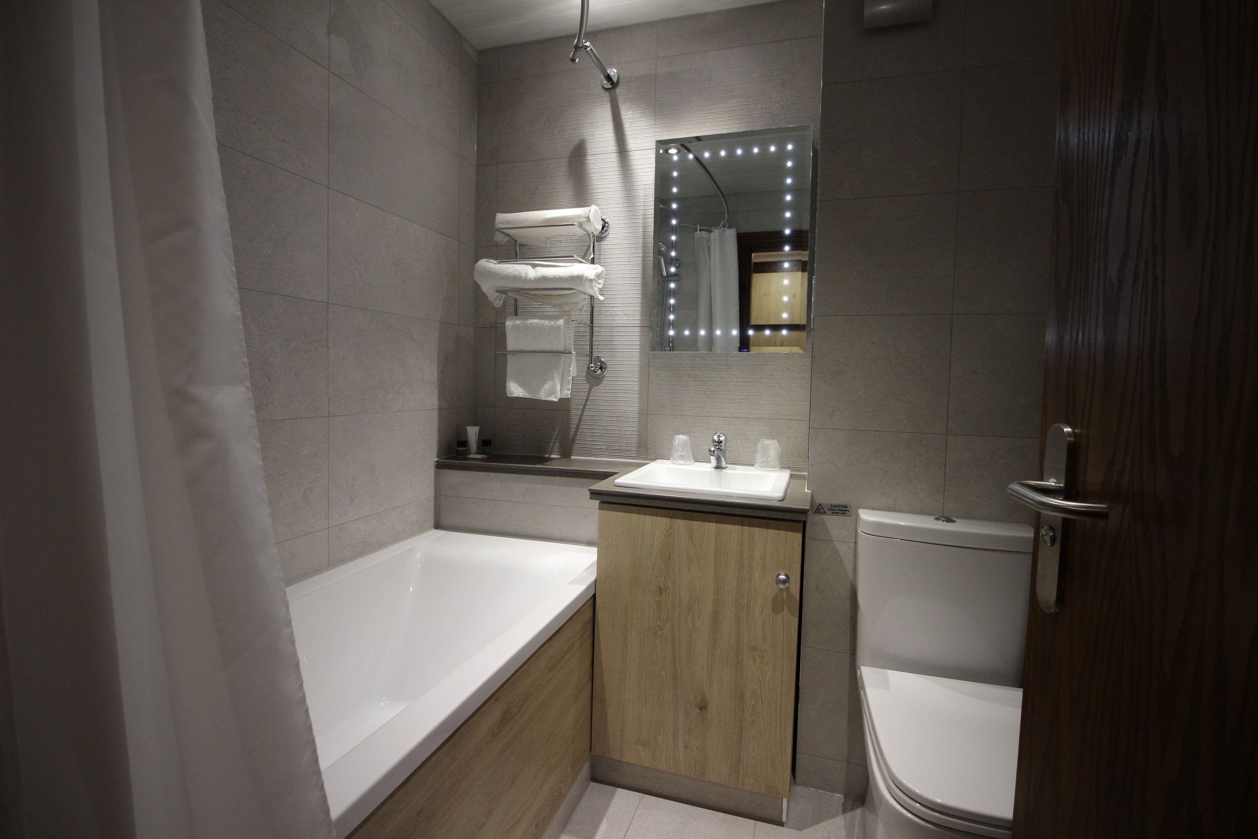 A modern bathroom with a bathtub, a sink, and a toilet. There is a vanity with a mirror and lights, and towels are hung on a rack.