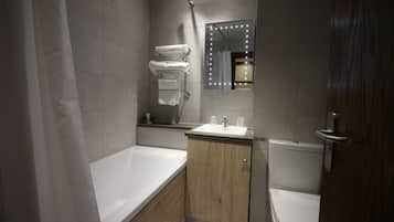 Un baño moderno con bañera, lavamanos e inodoro. Hay un mueble de tocador con espejo e iluminación, y toallas colgadas en un perchero.