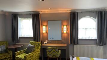 A hotel room with a bed, two chairs, a desk, a mirror, and two windows with curtains.
