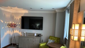 A hotel room with a flat-screen TV mounted on the wall, a minibar, a sofa with patterned cushions, a small round table, and a lamp with a patterned shade. - Pastures Hotel (Doncaster)