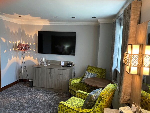 Ein Hotelzimmer mit Flachbildfernseher an der Wand, einer Minibar, einer Couch mit gemusterten Kissen, einem kleinen runden Tisch und einer Lampe mit einem gemusterten Lampenschirm.