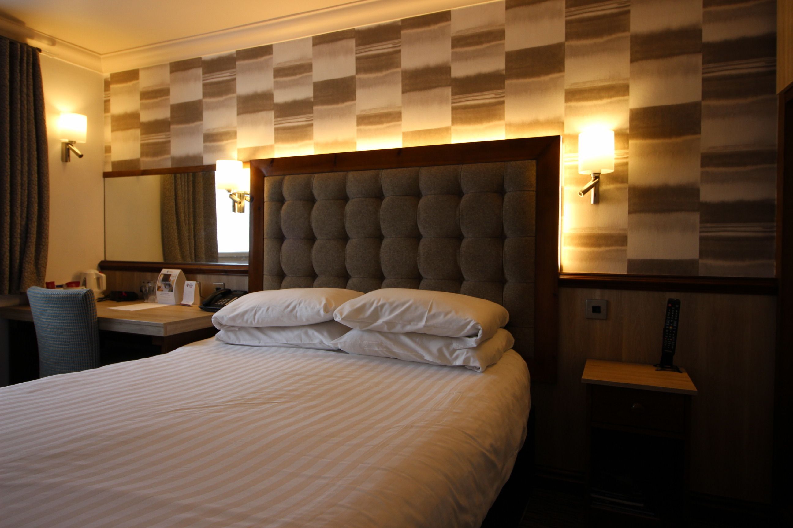 Ein Hotelzimmer mit einem großen Bett, einem Nachttisch, einem Sessel, einem Schreibtisch, einem Spiegel und Wandleuchten.