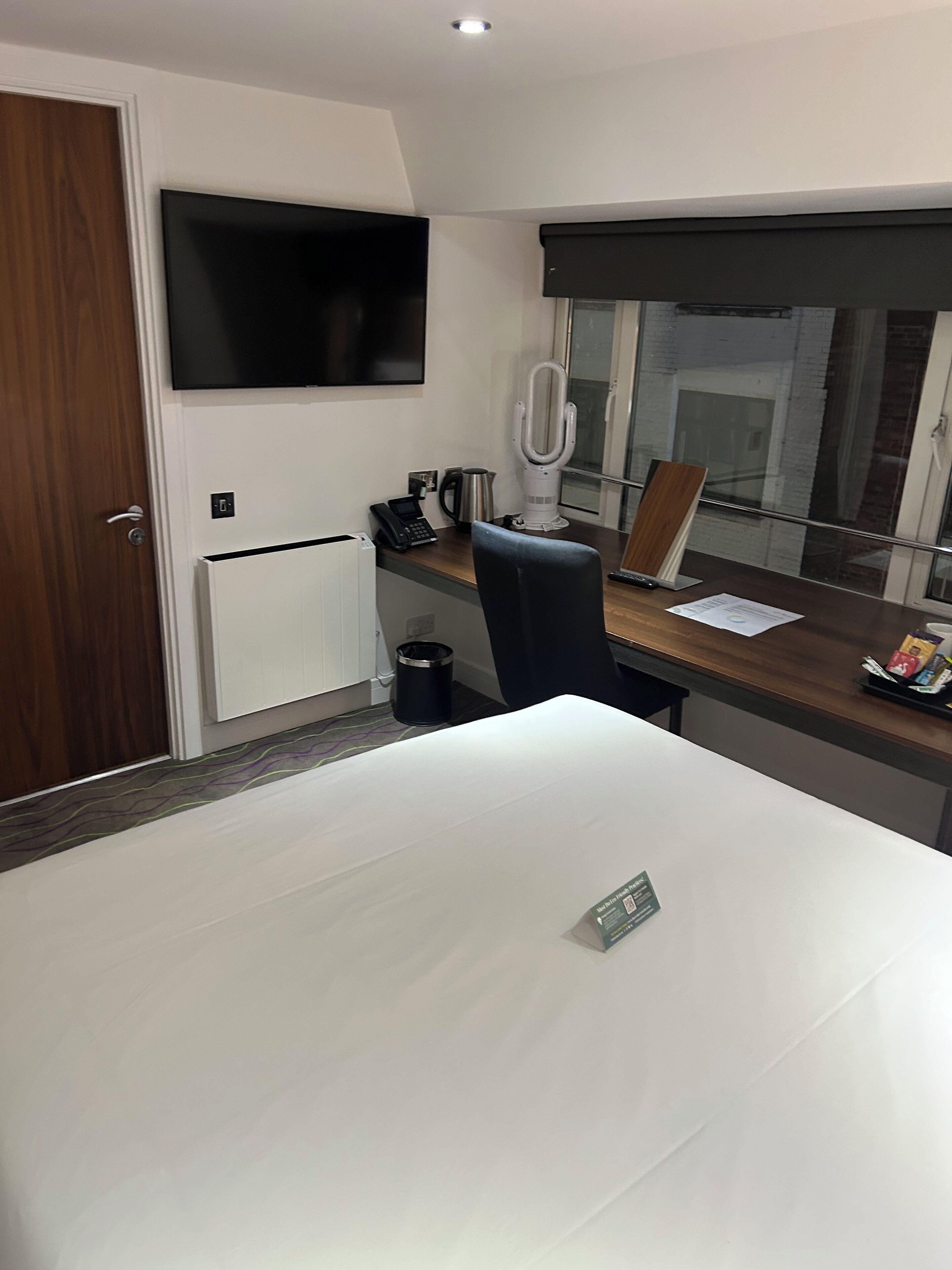 A hotel room with a large bed, a desk with a chair, a television mounted on the wall, a window with a view of the city, and a door with a wooden frame.