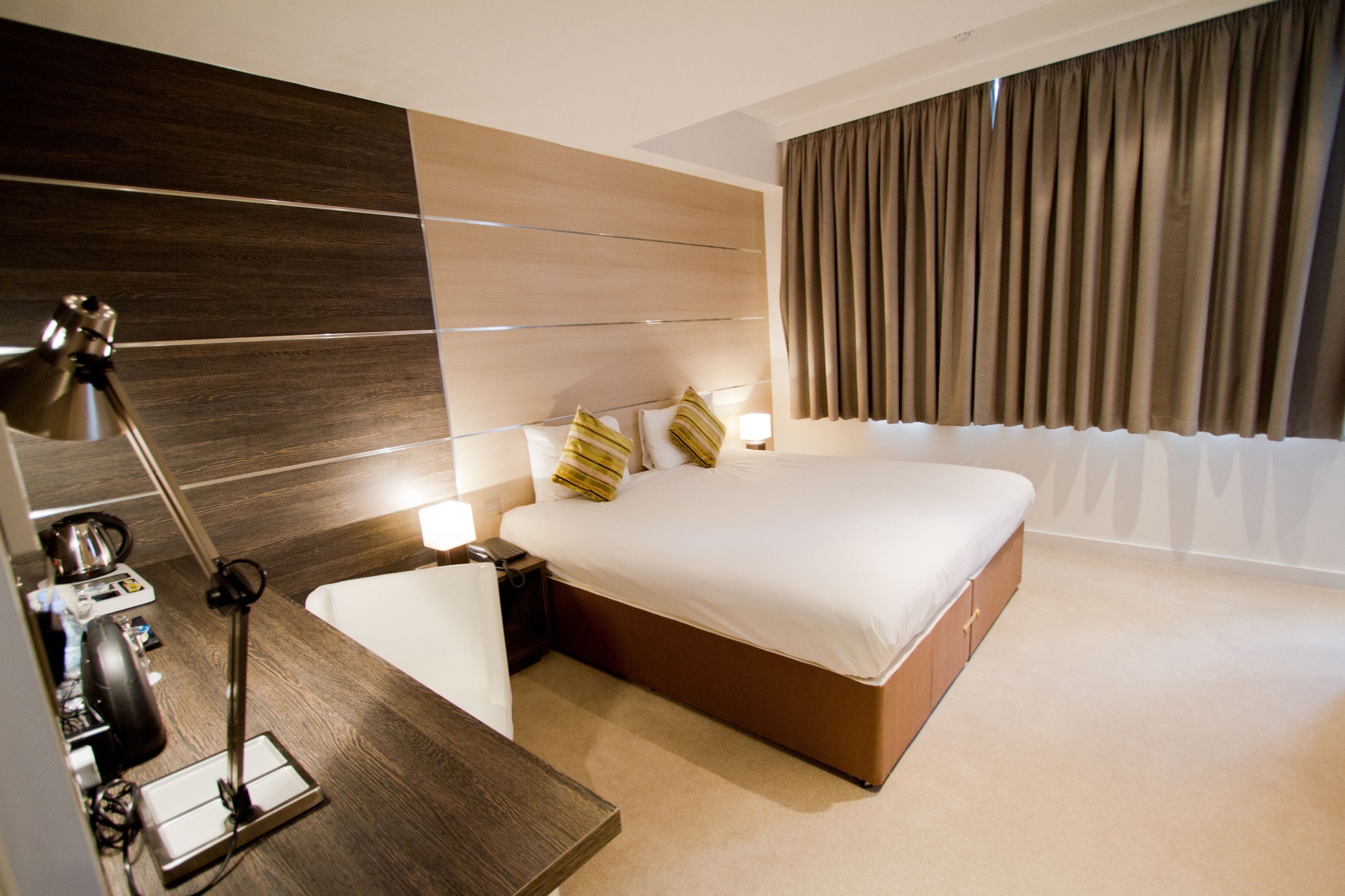 A modern hotel room with a large bed, bedside tables, a desk with a lamp, and a view of the outside through curtains.