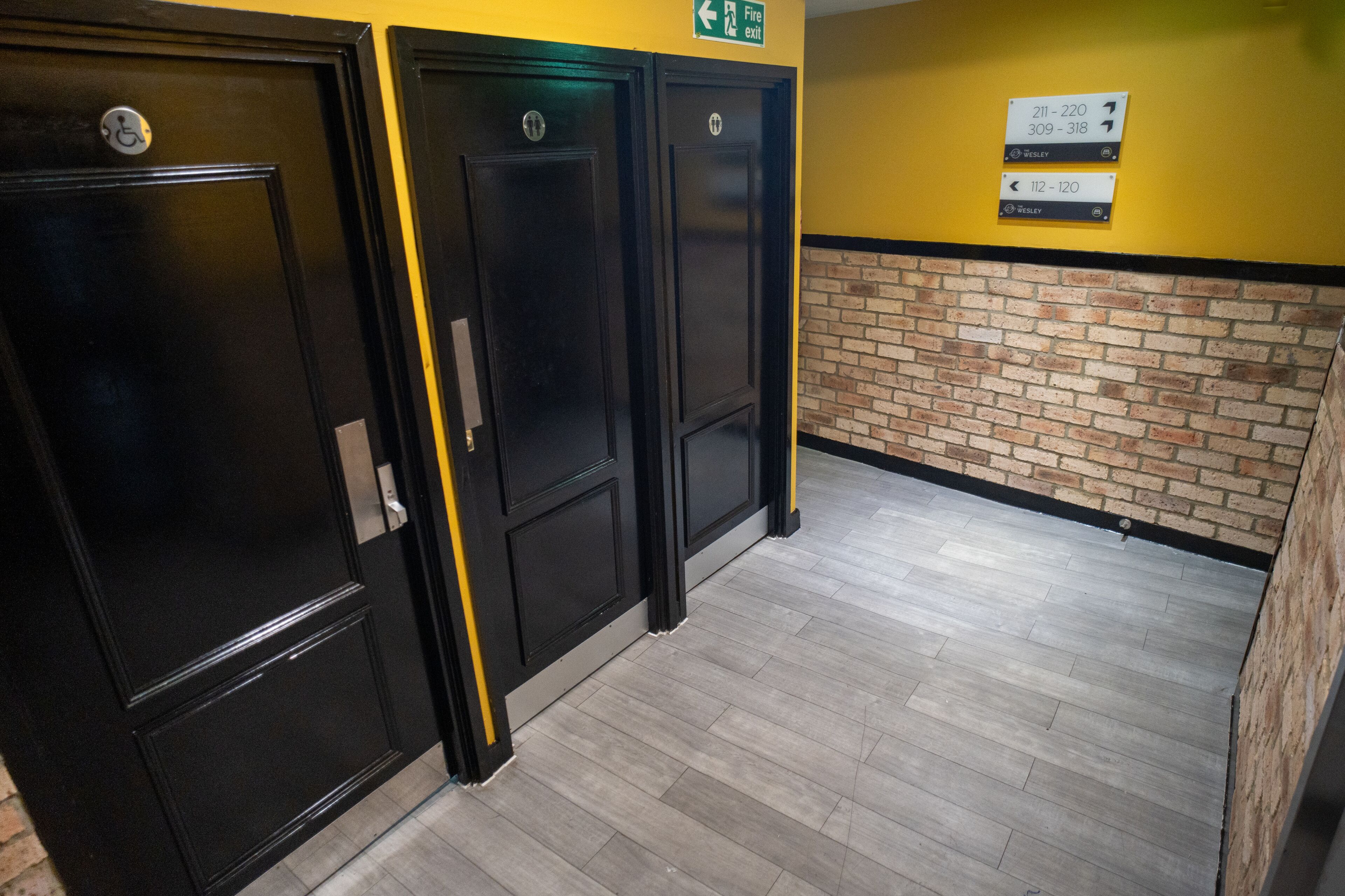 A hallway with black doors, a yellow wall, and a brick accent wall. There are signs indicating room numbers and directions.