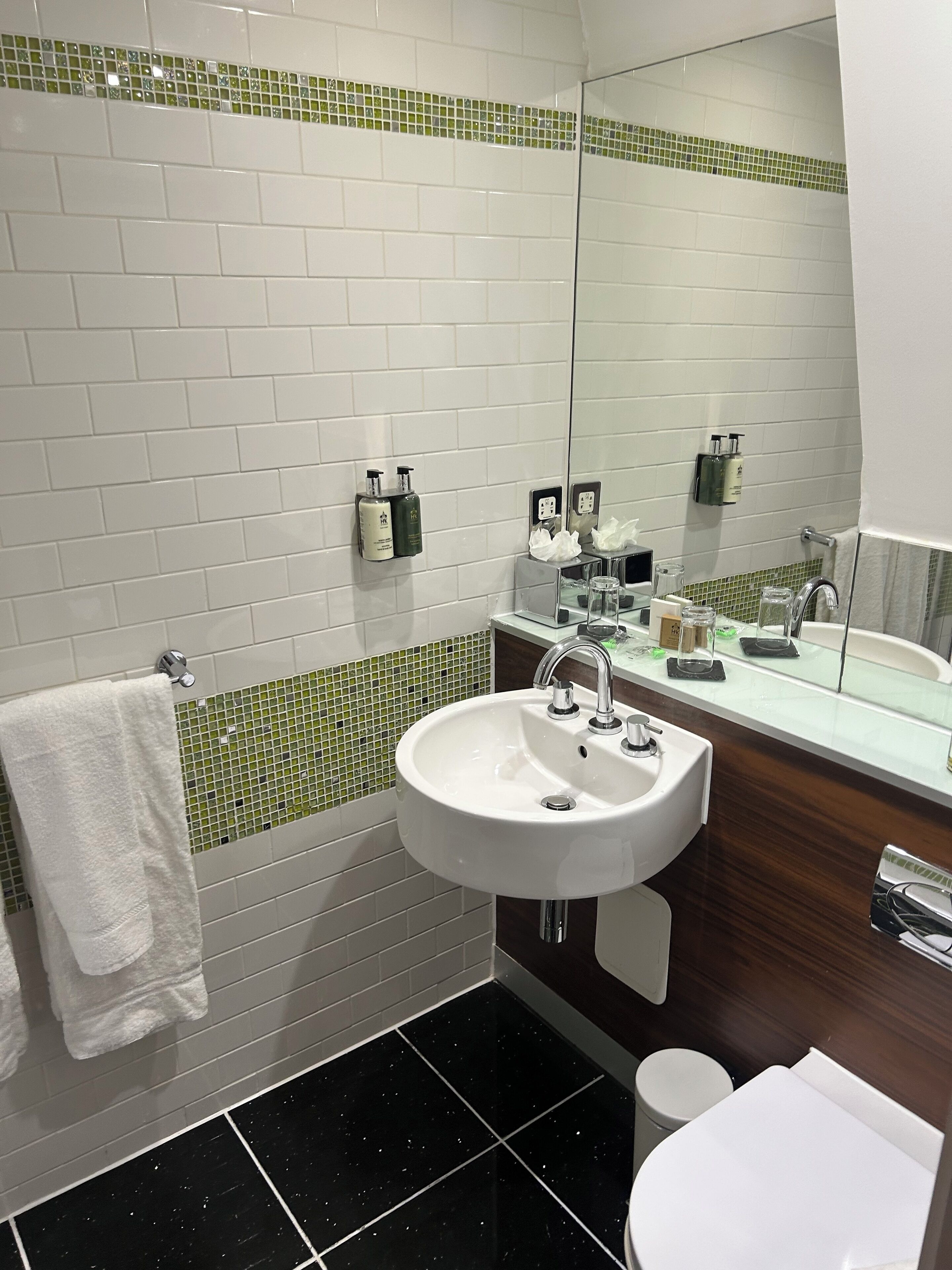 Un baño moderno con baldosas blancas tipo metro, un borde de mosaico verde, un lavamanos blanco con espejo y un inodoro.