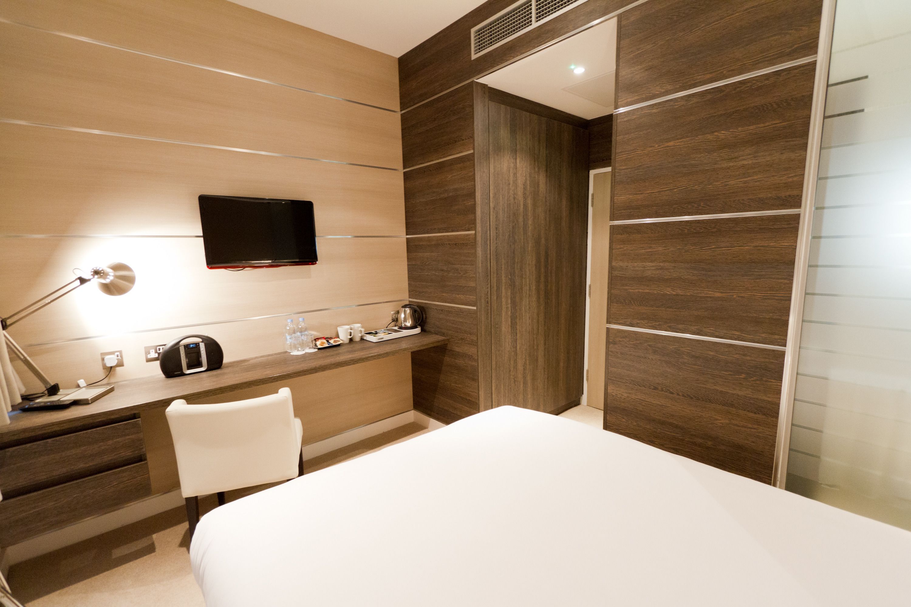 A modern hotel room with a bed, a desk with a lamp, a TV, and a minibar.