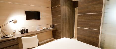 A modern hotel room with a bed, a desk with a lamp, a TV, and a minibar.
