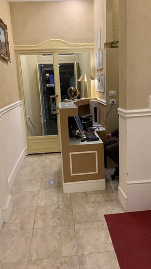 Een hotel lobby met een receptiebalie, een glazen deur die naar een kamer leidt, en een rood tapijt.