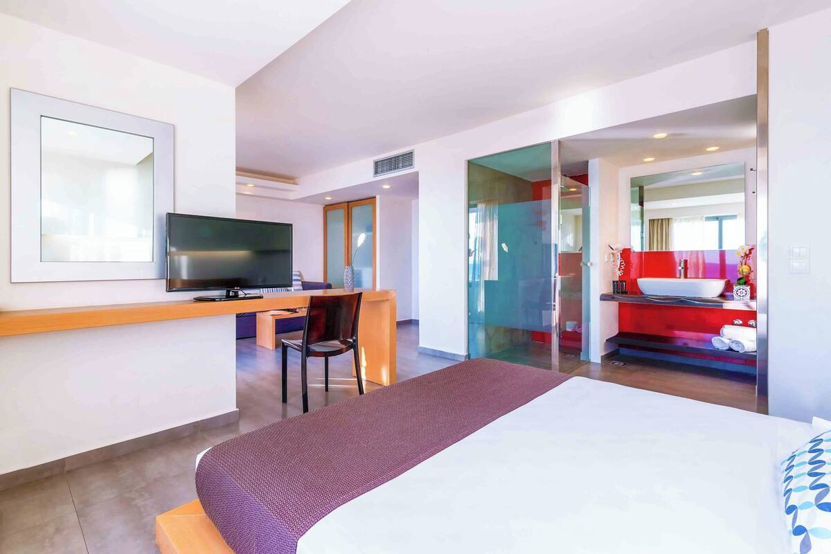 Habitación de hotel con cama, escritorio con televisor, una silla y un baño visible a través de una puerta abierta.