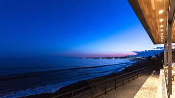 Vista costera desde un balcón con barandilla, con vista al mar y un paisaje urbano distante al anochecer.